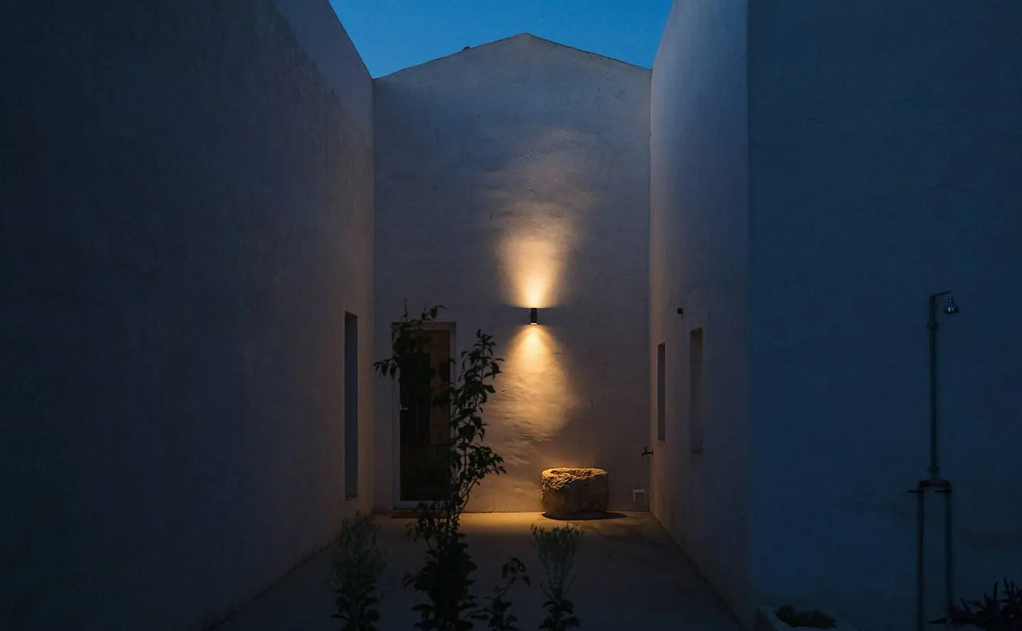 The narrow courtyard at blue hour, one lamp throwing light up the white wall