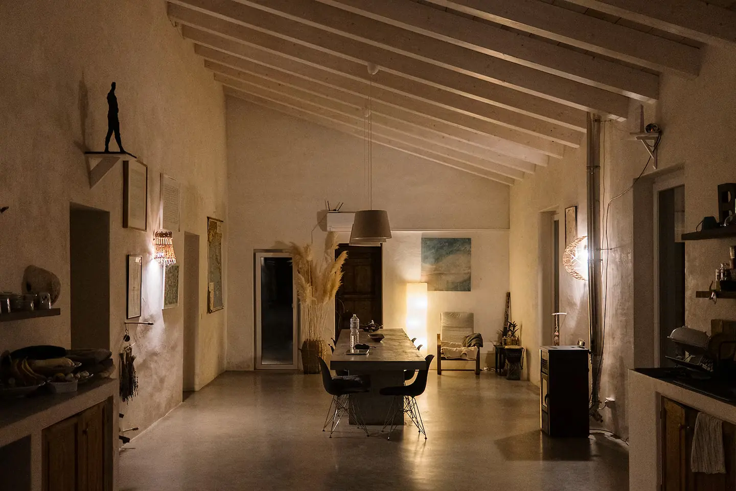 Inside at dusk, the long room lit by lamps with the dining table at the far end
