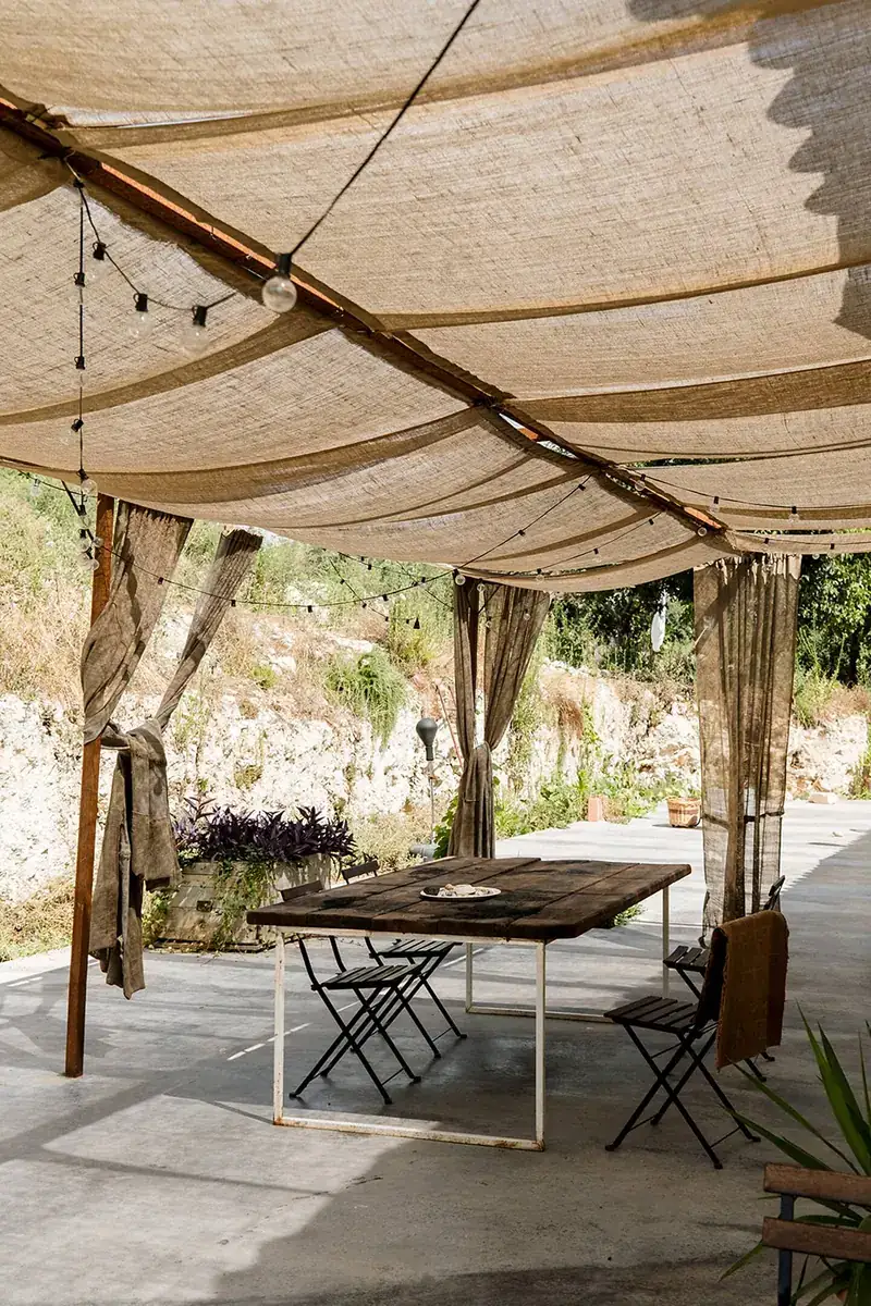 A long wooden table under the pergola, festoon lights strung through the draped canvas above